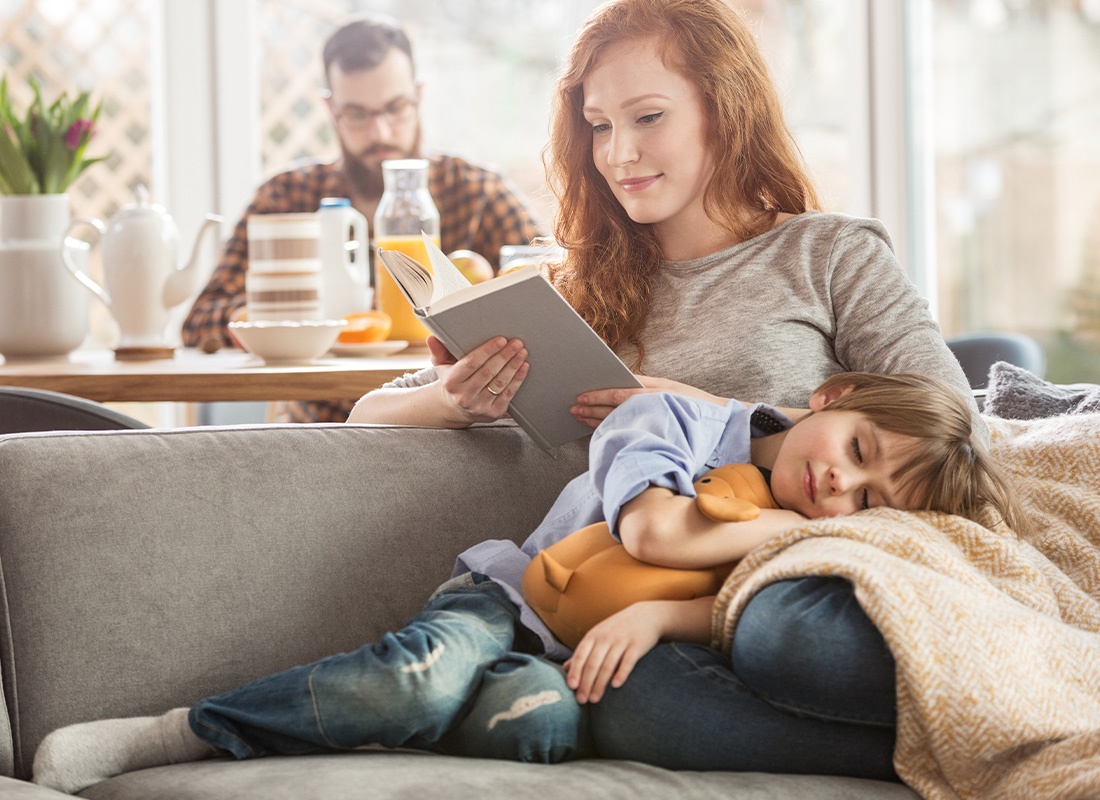 Personal Insurance - Young Son Sleeping On Mother’s Lap as She Reads on the Sofa With Dad in the Background